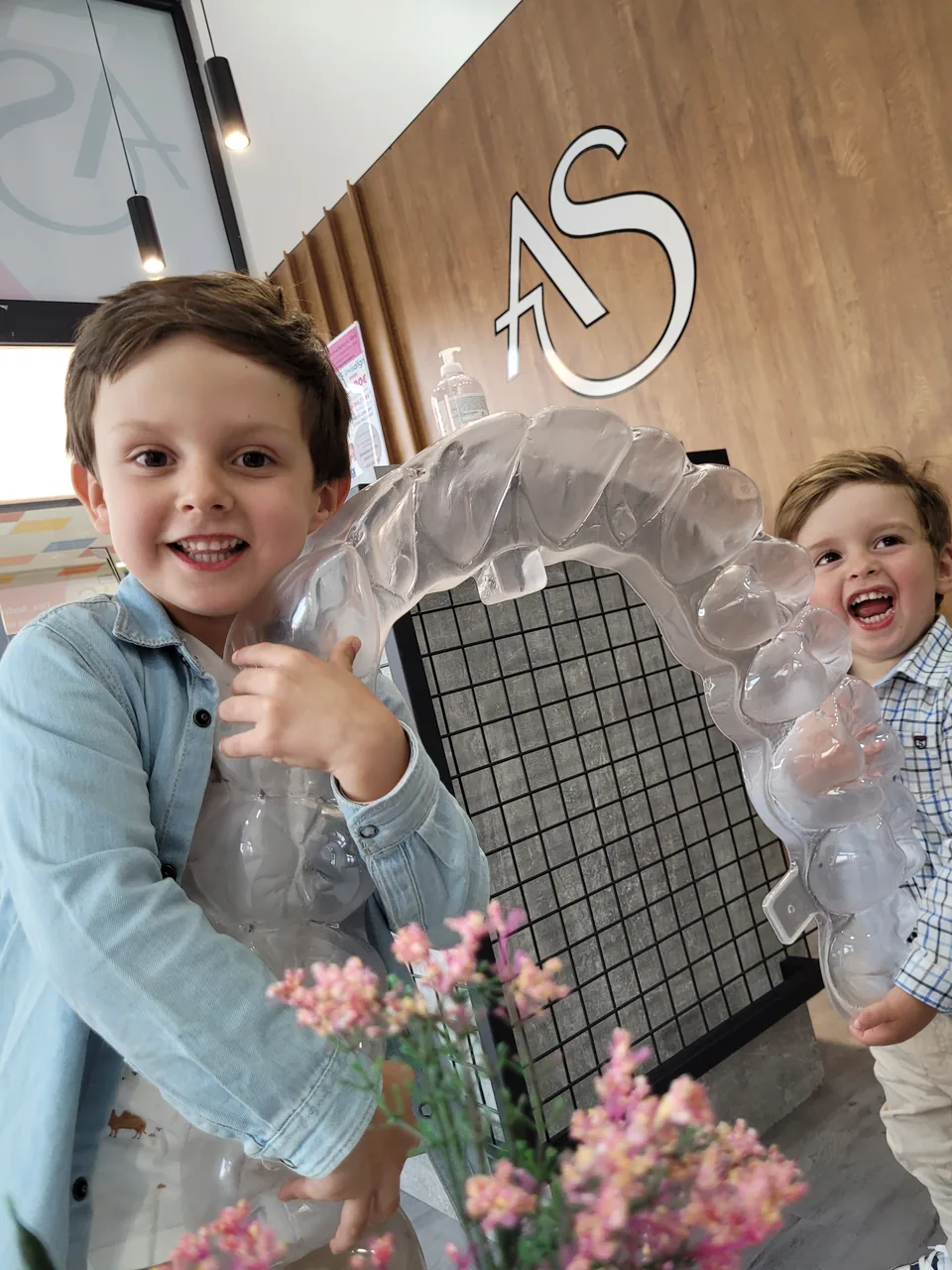 Dos niños sonriendo en la recepción de Ortodoncia Santonja sujetando un modelo de alineador, bajo el logotipo AS